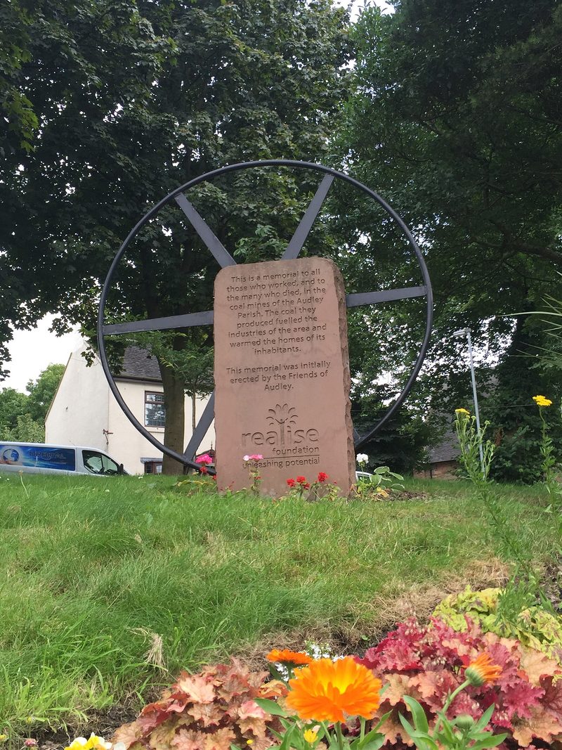 A stone mining memorial in Audley village with a plaque and a pit wheel, surrounded by paving and planting