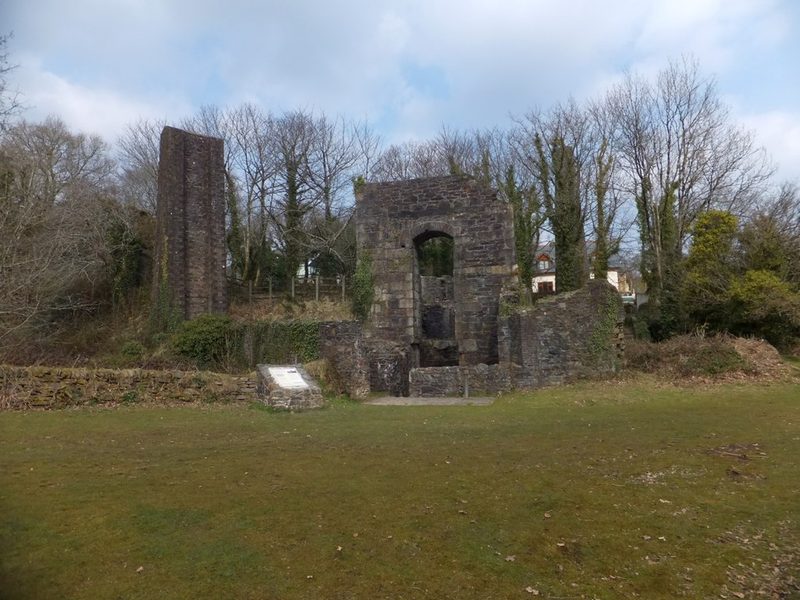 A roofless stone engine house with an arched opening and a tall stone stack beside it, behind a grassy lawn.