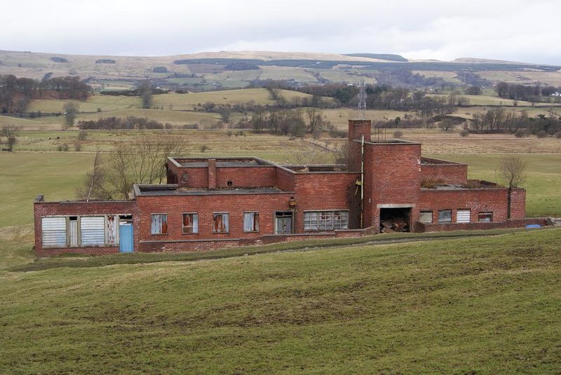 A long flat-roofed red brick building with boarded windows and a small tower, standing in a grassy field below hills.