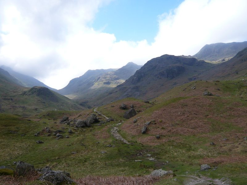 A path winding up a green valley towards the rocky knoll of Eagle Crag, with the high Helvellyn fells behind under cloud.
