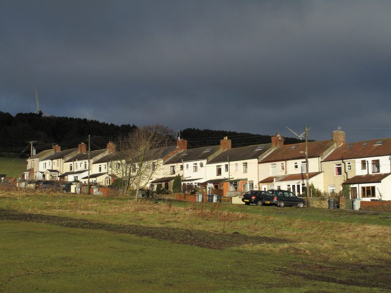 A long terrace of pale colliery cottages behind rough grass under a dark sky, with wooded hills behind.
