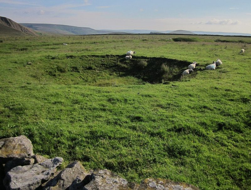 A grass-covered, roughly circular collapsed mine shaft depression in an open Peak District field, sheep grazing around its rim, a drystone wall in the foreground and hills on the horizon.