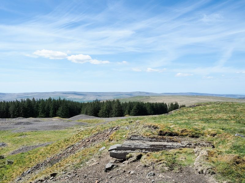 A grassed mound of mine spoil on open moorland with concrete railway sleepers embedded in its top.