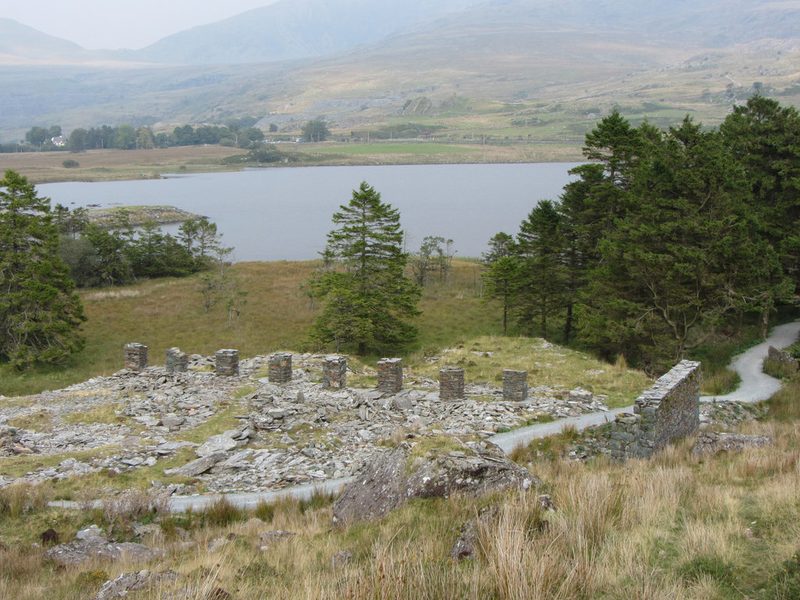 A row of stone pillars stands among slate waste beside a path, with a lake, conifers and mountains beyond.