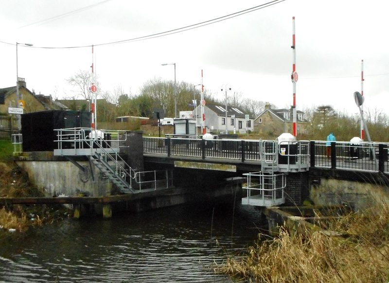 A stone-and-steel road bridge over a canal in a wooded cutting, with towpath and moored water below.