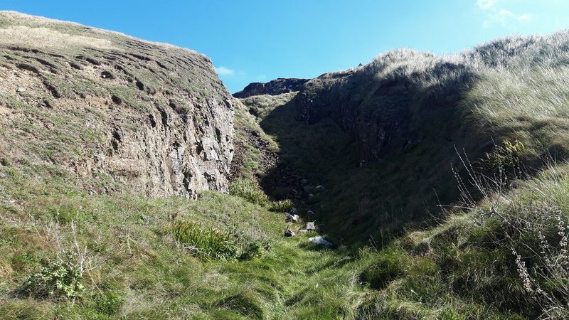 A steep-sided open cut through grass-covered cliff-top ground at Gravel Hill, with bare rock faces on either side where the iron lode was worked.
