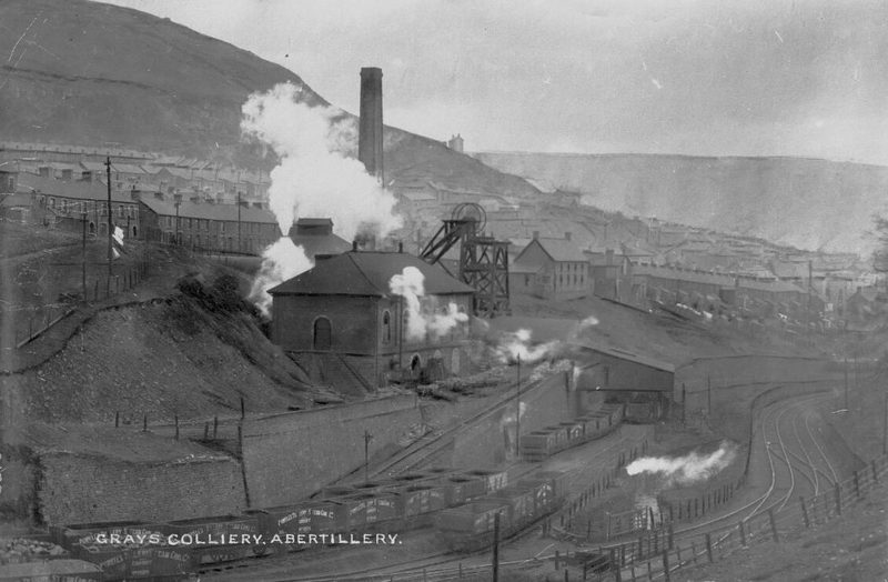 Black and white view of a colliery with a headframe, engine house and tall chimney below terraced streets on a hillside.