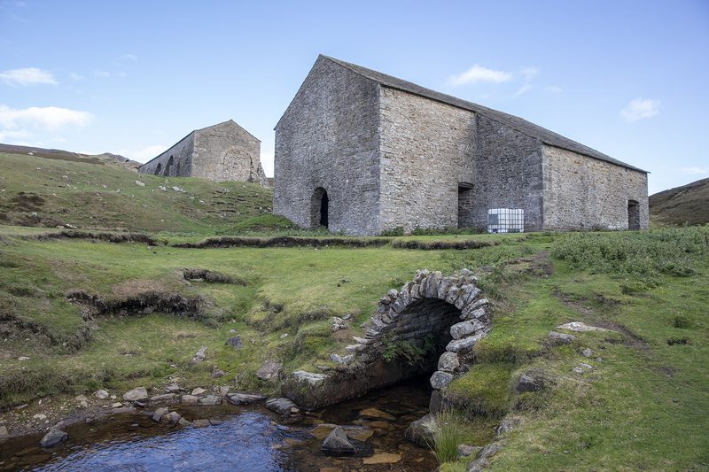 Two roofed stone buildings on a moorland slope, with a small stone-arched culvert over a stream in the foreground.