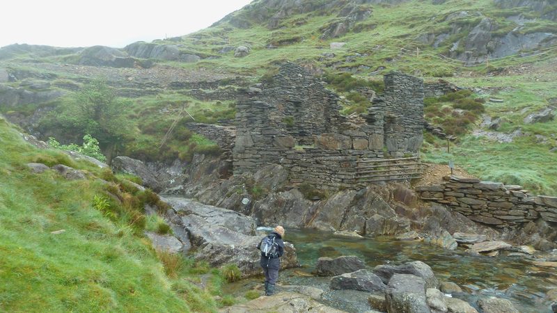 A walker stands by a mountain stream below a tall roofless slate-built ruin on a grassy hillside.