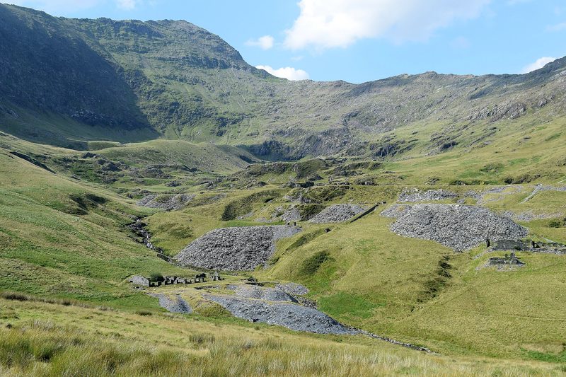 A green mountain valley below a rocky ridge, with grey slate tips and roofless quarry buildings on its floor.
