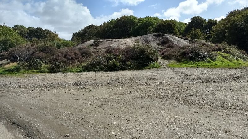 A bare grey spoil heap of mine waste, partly overgrown with heather and scrub, beyond a gravel track at Halamanning in 2017.
