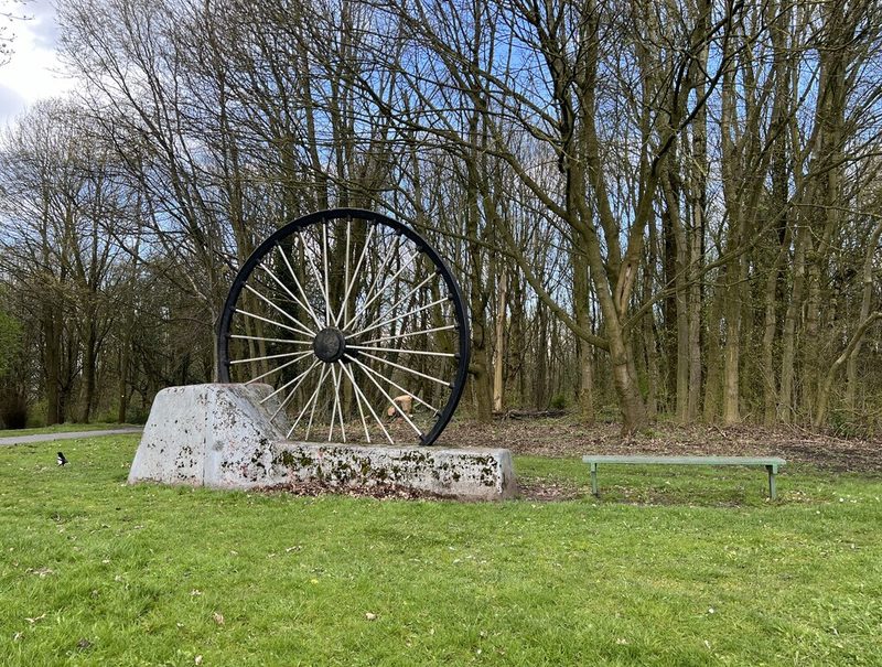 A large black pithead wheel mounted on a concrete plinth in parkland among bare trees.