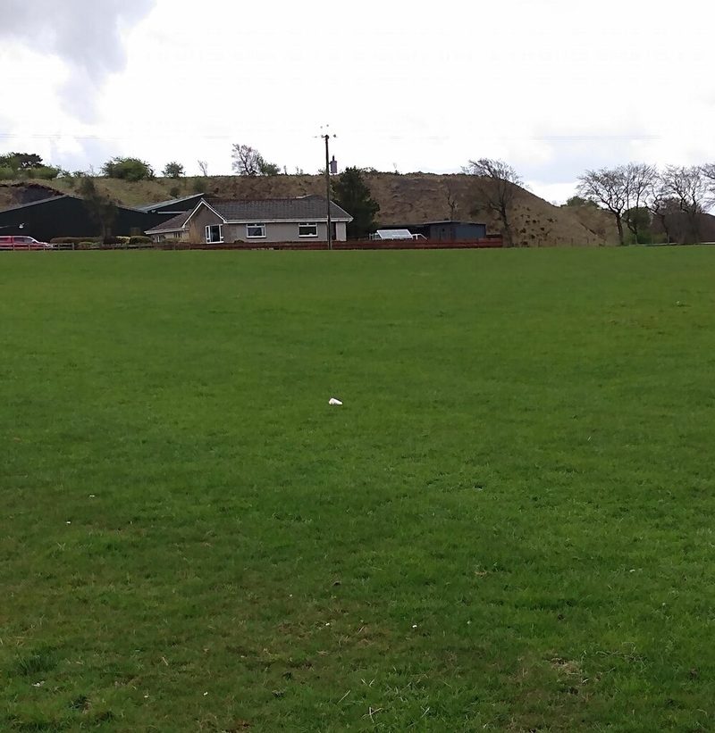 A bungalow and farm sheds beyond a large grass field, with a long brown spoil heap rising behind them.