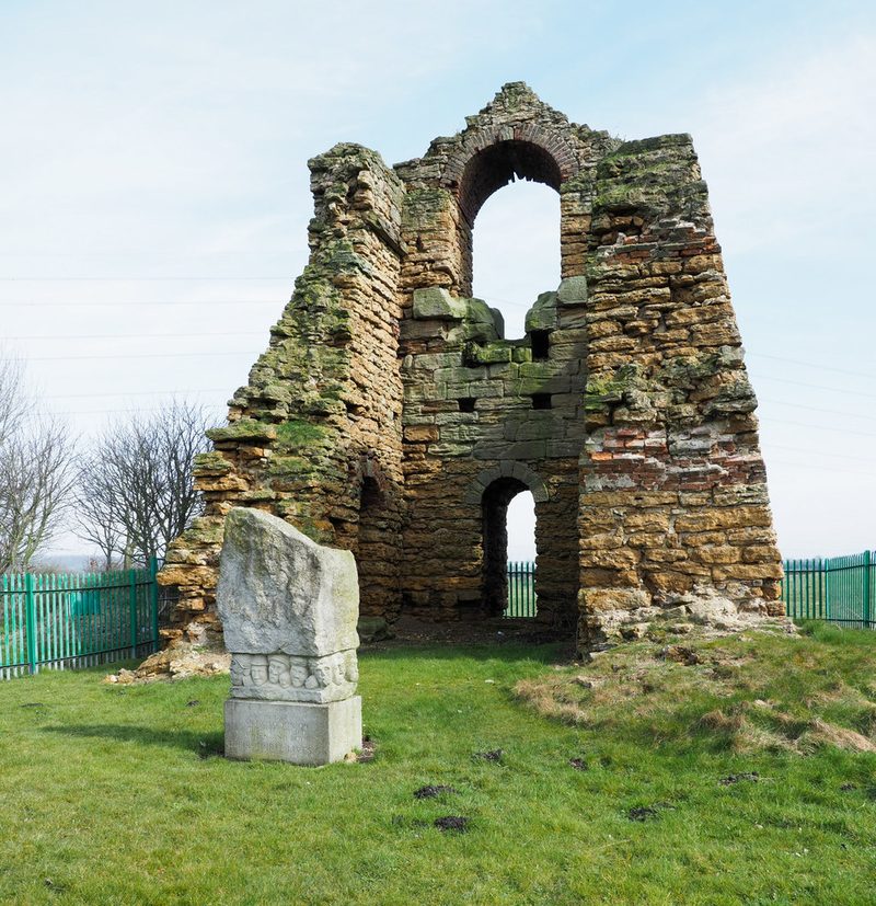 The roofless stone shell of a tall engine house with an arched upper opening, a carved memorial stone of faces in front, inside green railings, 2018