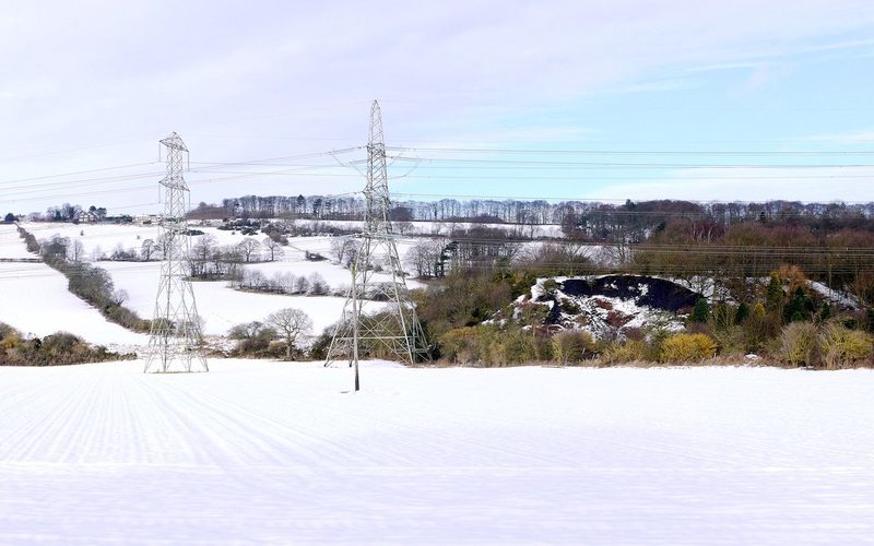 A dark spoil heap among scrub in a snow-covered valley, with electricity pylons in the foreground.