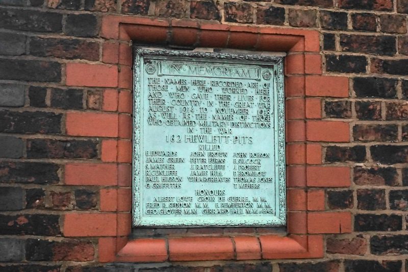 A weathered bronze plaque headed In Memoriam and listing names, set in a brick wall with a red-brick surround.