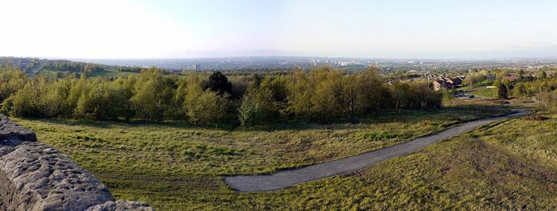 A grassy hilltop path winding through young trees, with a wide view over Tyneside towns.