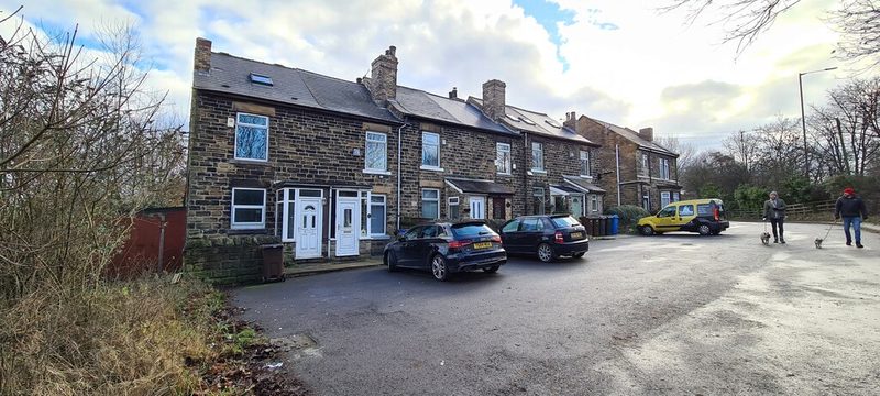 A terrace of red-brick cottages beside a road lined with parked cars under a grey sky.