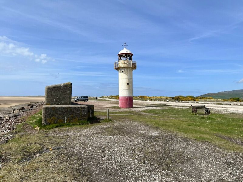 A short white and red iron lighthouse on level gravelly ground beside the sea wall, with a concrete block and a bench in front.