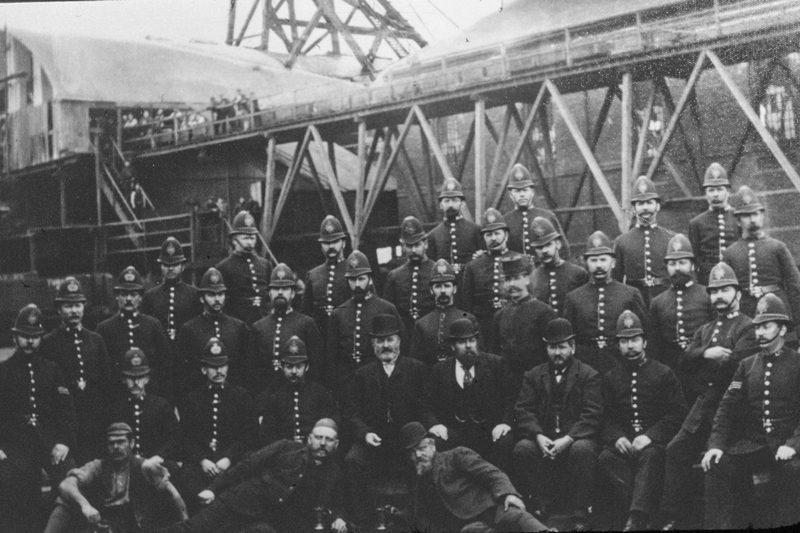 Rows of helmeted policemen and three men in bowler hats pose in front of a timber headgear and gantry.
