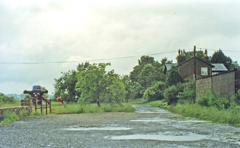 A wet gravel track runs past a stone house and a hedged field where the railway once ran, under a grey sky.