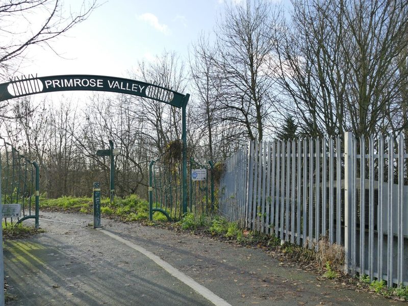 A path passing under a green arched Primrose Valley sign beside a tall steel palisade fence.