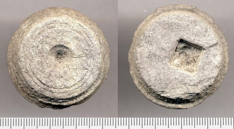 A flat circular disc of black shale with a square recess cut off-centre in one face, the waste core left in a lathe after an armlet was turned from it.