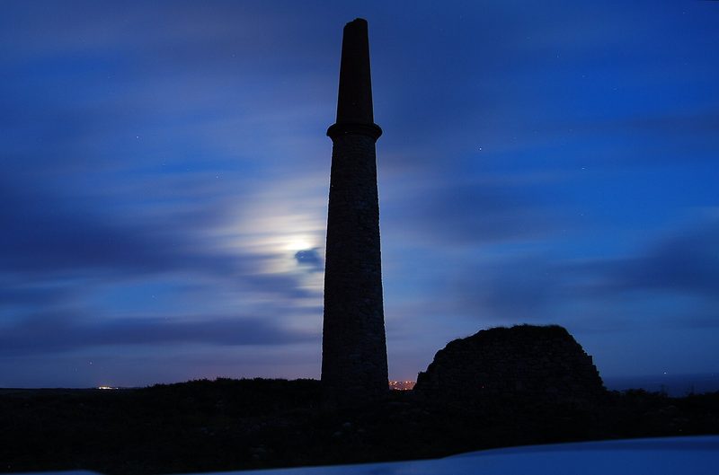 A tall stone chimney and a ruined wall stand silhouetted against a moonlit blue night sky.