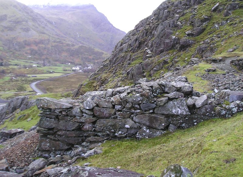 A drystone retaining wall on a grassy slope below crags, with a road winding up a valley beyond.