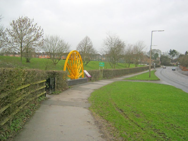 A yellow-painted colliery winding wheel standing as a memorial beside a footpath and a main road.
