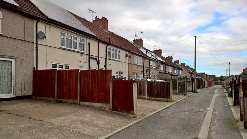 A straight street of terraced brick cottages with small front gardens and parked cars