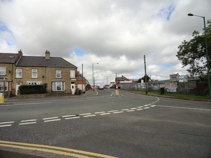 A road junction with stone terraced houses on one side and a tyre yard behind railings on the other