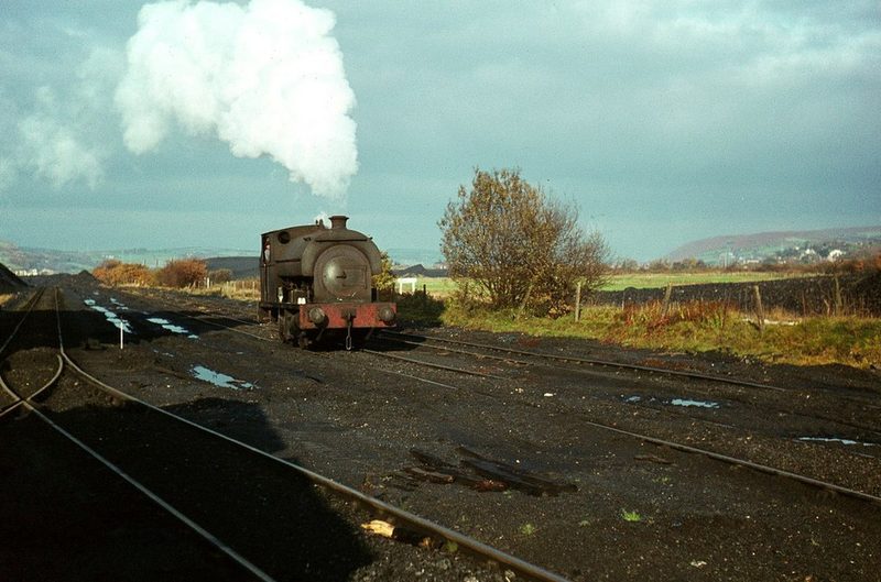 A small steam saddle-tank locomotive puffing white smoke on coal-blackened sidings under a pale sky.
