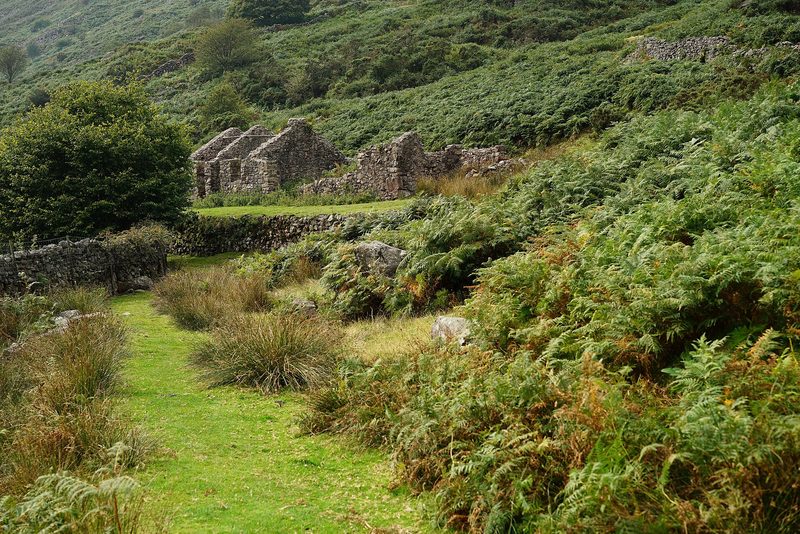 Roofless stone buildings on a grassy platform at the foot of a bracken-covered fellside, with a path in the foreground.