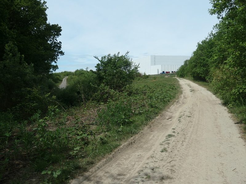 A sandy track runs between bushes and trees towards a large white warehouse on the former colliery site.