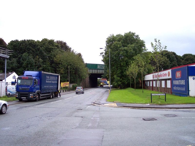 A road under a railway bridge lined by trees, with a lorry and warehouses beside it.