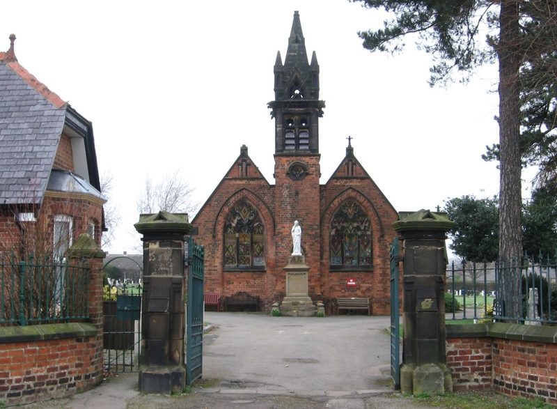 Cemetery gates opening onto a brick chapel with a white memorial statue on a pedestal in front.