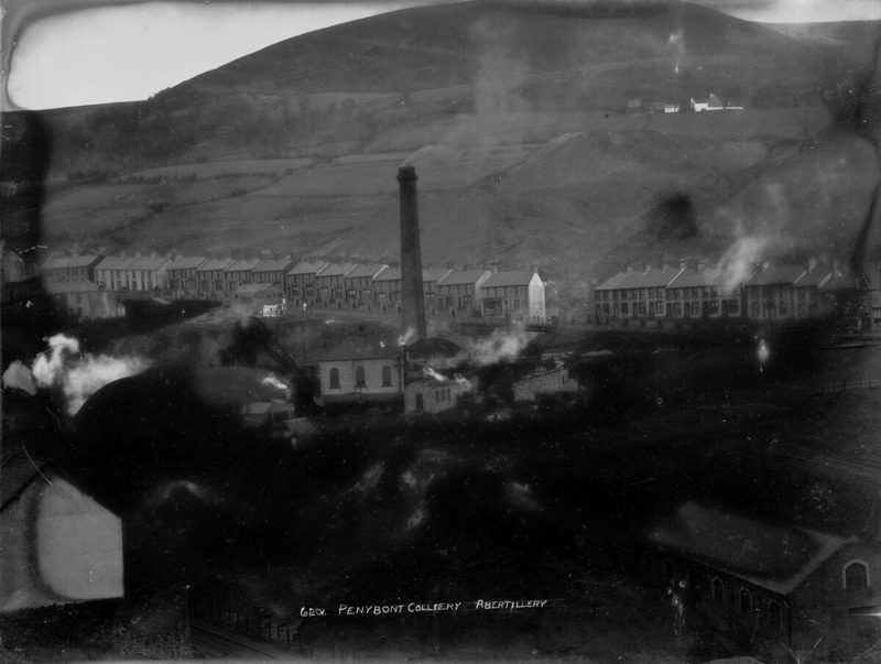 Black and white view of a colliery chimney and engine houses below rows of terraced houses on a steep hillside.