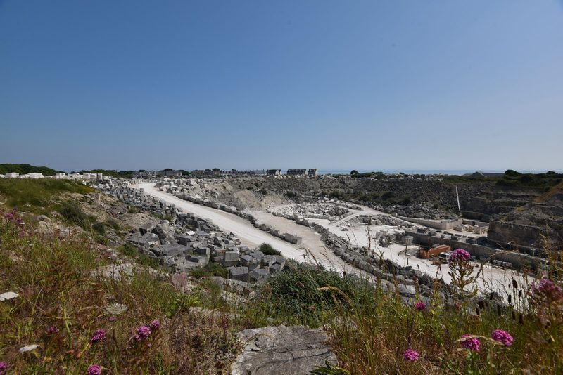A wide working quarry with rows of cut pale stone blocks along white tracks, houses on the skyline and the sea beyond.