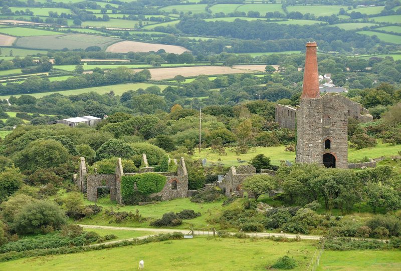 Stone engine house with a tall square-based chimney topped in red brick, beside roofless ruins of mine buildings among trees and fields.