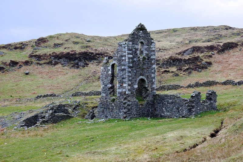 A tall ruined stone engine house with arched openings on a rough grassy hillside, with lower walls beside it.