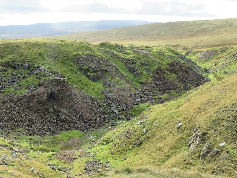 A deep grassed gully with rocky spoil banks cut into the moorland, the hushed ground falling away towards distant fells.