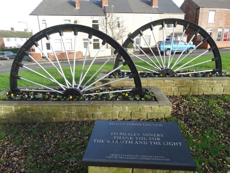 Two black pit wheels set in stone flower beds behind a slate plaque reading To Pilsley miners, thank you for the warmth and the light.