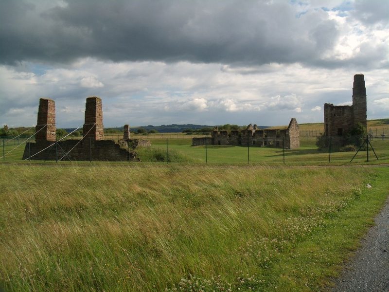 Ruined stone chimney stacks and roofless buildings behind a wire fence on a grassy site under a cloudy sky.