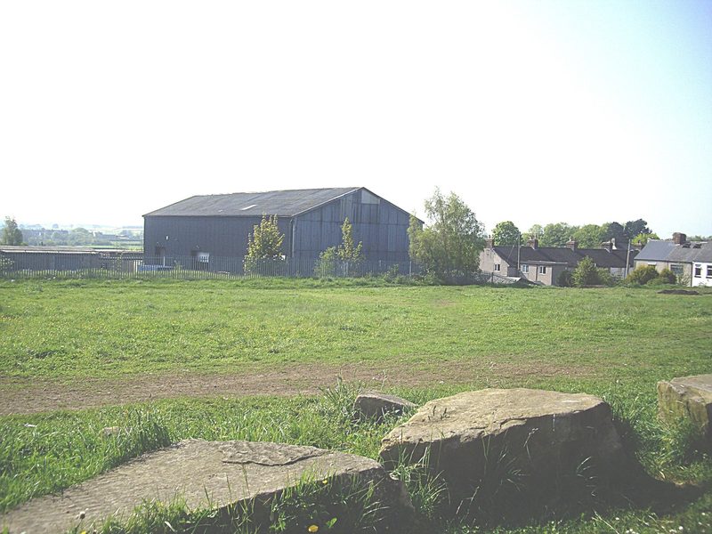 A grassy open field with large stone blocks in the foreground, a corrugated industrial shed and a terrace of houses beyond.
