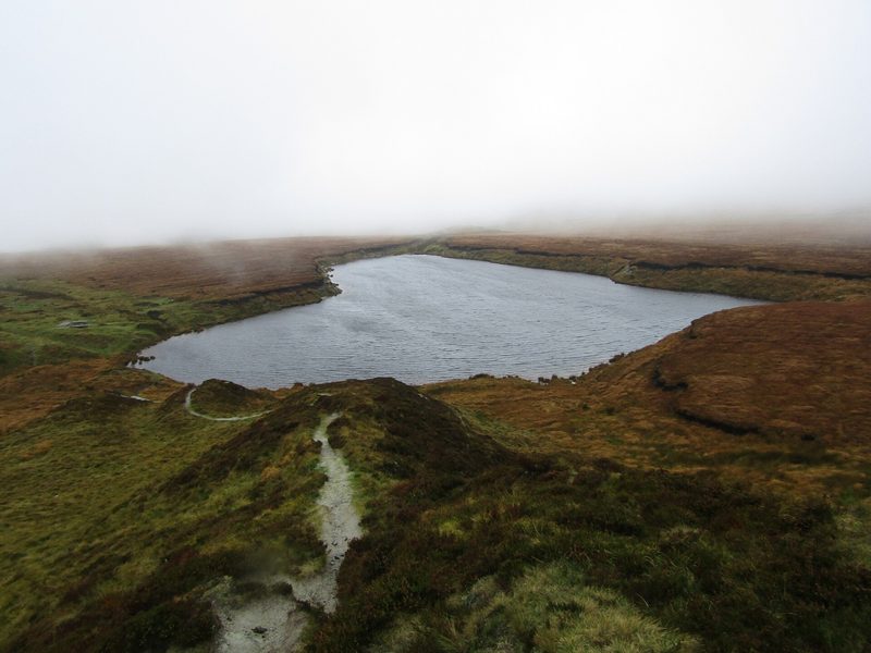 A grey water-filled pit in brown moorland seen from a heathery ridge, with low mist over the hills.