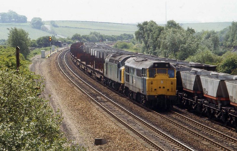 Two diesel locomotives hauling a steel train past lines of coal hopper wagons in the colliery sidings