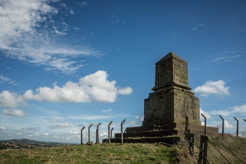 The squat stone base of a monument standing on the grassy summit of Bignall Hill behind a wire fence, with open country beyond.