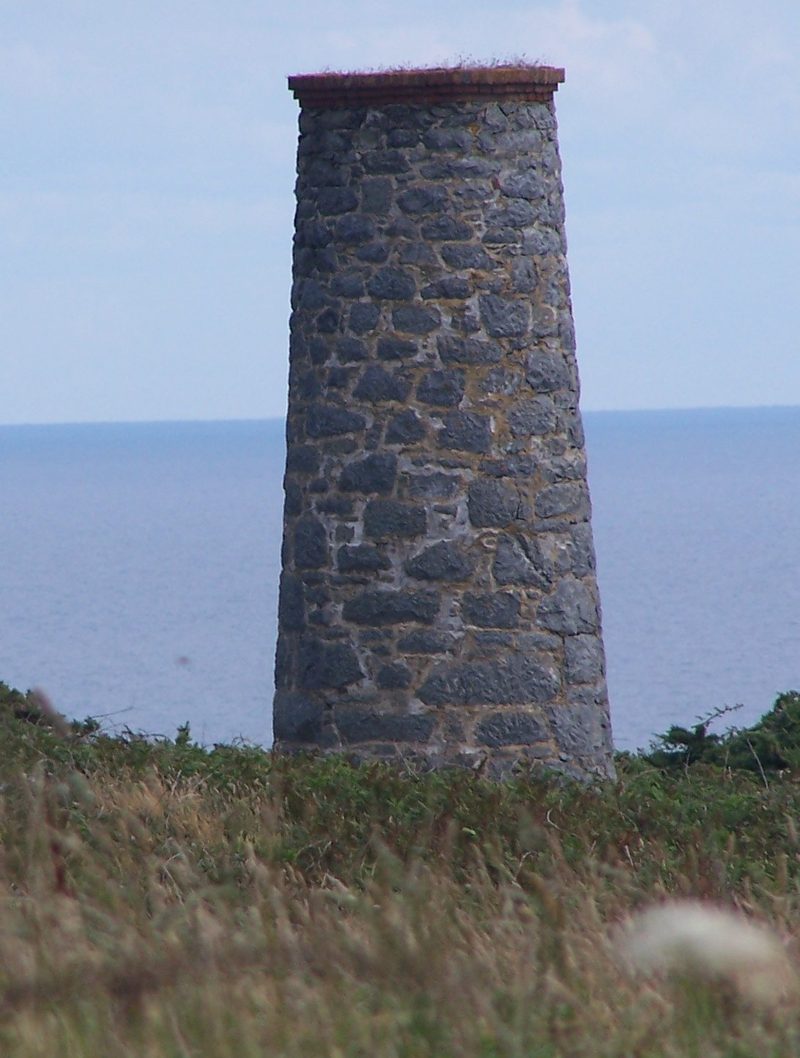 A tapering stone chimney stack standing alone in rough grass on a clifftop, with the sea behind it.