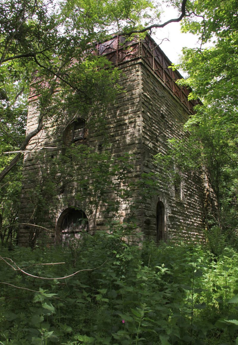 A tall sandstone engine house with arched openings and a rusted iron water tank on top, half hidden by trees and nettles.